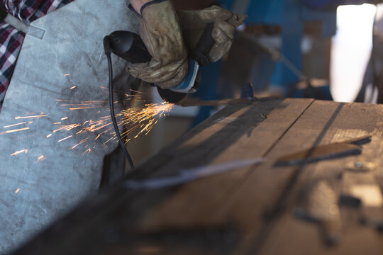 Hands of caucasian male knife maker wearing apron, using angle grinder in workshop - Powered by Adobe
