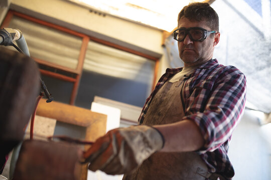 Caucasian male knife maker wearing apron and glasses, making knife in workshop