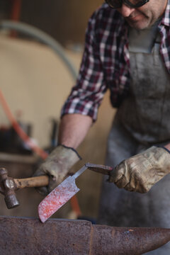 Close up of caucasian male knife maker wearing apron and glasses, using hammer in workshop