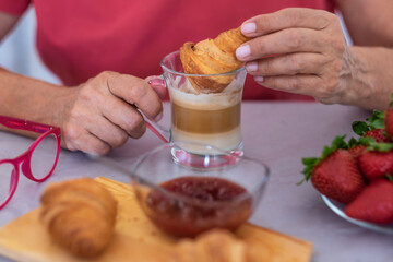 Close up on human hand of mature woman enjoying breakfast at home with fresh croissant and cappuccino. Strawberries and jam on the table