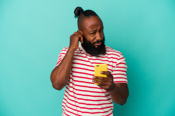 African american man holding a mobile phone isolated on blue background covering ears with hands.
