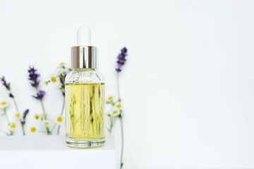 glass cosmetic bottle for serum or oil on white podium with lavender and chamomile flowers. Aromatherapy and spa cosmetic product concept. Copy space