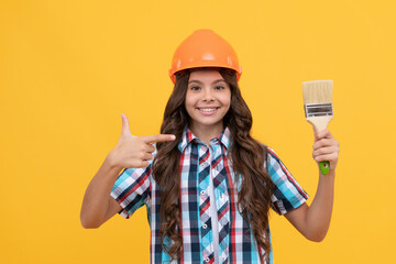 happy child with curly hair in construction helmet pointing finger on painting brush, renovation