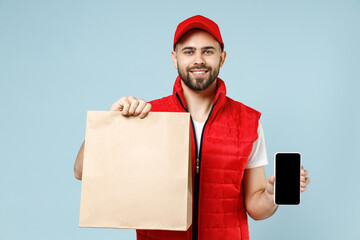 Delivery guy employee man in red cap white T-shirt vest uniform work as dealer courier service hold brown clear blank craft paper takeaway bag use cell phone isolated on pastel blue background studio.