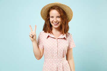 Smiling happy young redhead curly woman 20s show victory sign wears casual pink dress straw hat look camera isolated on pastel blue color background studio portrait. People emotions lifestyle concept.