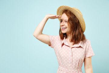 Smiling cute fun happy young redhead curly woman 20s years old wears casual pink dress straw hat hold hand at forehead look far away distance isolated on pastel blue color background studio portrait.