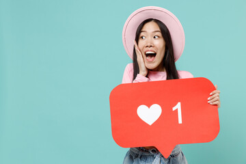 Fun young brunette asian woman 20s with open mouth wear pink clothes touch cheek look aside hold huge like sign from social network heart form isolated on pastel blue color background studio portrait