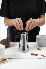 Cropped shot: lady in black is making coffee and breaking cinnamon stick above stainless steel moka pot. Glass jar with ground coffee and white cups are located near on the white table. 