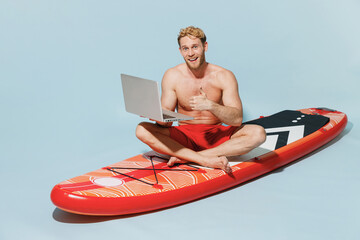 Full length young fun man in red shorts swimsuit sit on sup board use laprop pc computer work...