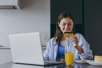 Young housewife woman in casual clothes blue shirt use laptop pc computer credit bank card shopping online order delivery book tour cook food in light kitchen at home. Healthy diet lifestyle concept.