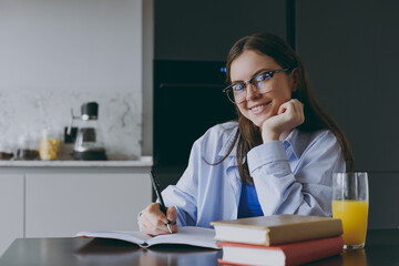 Smart smiling happy young housewife woman 20s in blue shirt casual clothes glasses writing down recipe in notebook diary prop up chin cook food in light kitchen at home Healthy diet lifestyle concept