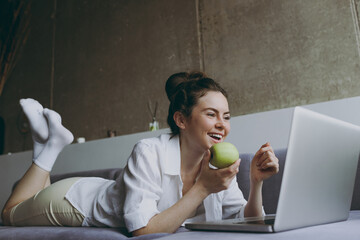 Full length young side view smiling freelancer woman in white clothes lying on grey sofa indoor...