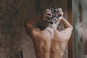 Back rear view young brunet half naked torso topless brunette man 20s taking hot shower wash hair with shampoo indoors in bathroom at home. People healthcare daily morning routine lifestyle concept