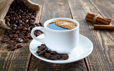Black coffee in a white cup, coffee beans, cinnamon sticks on an old wooden background.