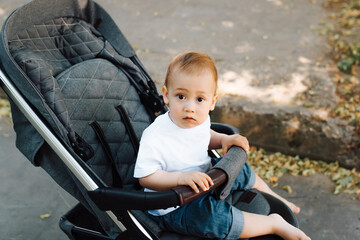 Portrait of a funny little child sitting in a stroller and looking at the camera, outdoors. Kid in a baby carriage for a walk on a summer day.