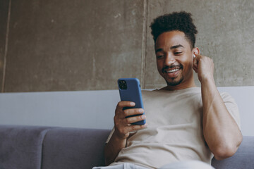 Young happy african american man in beige t-shirt sitting on grey sofa indoors apartment use air...