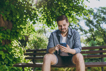 Fototapeta premium Bottom view smiling satisfied young man in blue shirt sit on bench use mobile cell phone chat online rest relax in spring green city sunshine park outdoors on nature Urban lifestyle leisure concept