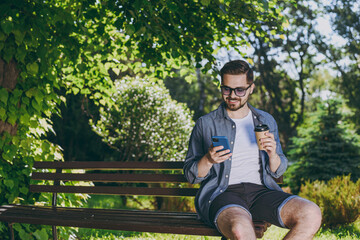Young man in blue shirt shorts glasses sit on bench hold mobile phone takeaway cup drink coffee rest relax in spring green city park sunshine lawn outside on nature Urban lifestyle leisure concept