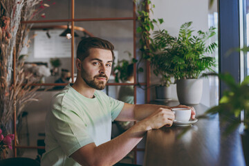 Side view young confident caucasian man 20s in mint casual t-shirt drink tea hold cup of coffee...