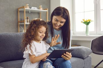 Happy young Caucasian mother and little daughter having fun with modern tablet device. Smiling mother and little girl waving hand greeting and talking on online video call on pad device.