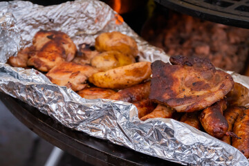 Process of cooking fresh meat steaks, sausages and potatoes in foil on brazier at summer local food market - close up view. Outdoor cooking, gastronomy, cookery, street food concept