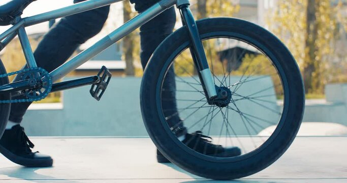 Teenager in black tight pants, sneakers walks confidently down ramp at skatepark, trees in background, bike in foreground, spinning wheels, bmx spokes, fat tire, low frame