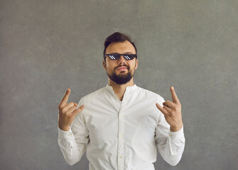 Self assured man wearing funny sunglasses. Studio shot of smirking smug looking student or businessman in white shirt and thug life glasses doing rock sign gesture standing isolated on grey background - obrazy, fototapety, plakaty