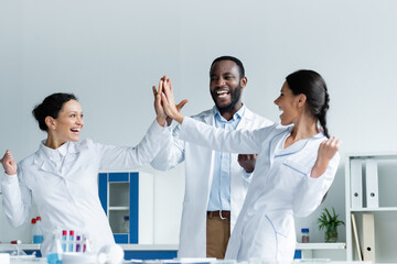 Cheerful multiethnic scientists giving high five near medical equipment in lab - Powered by Adobe
