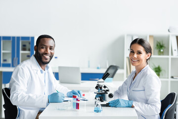 Fototapeta premium Smiling multiethnic scientists looking at camera near medical equipment