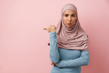 A sad muslim woman wearing pink hijab pointing to the copyspace and wrinkling her forehead