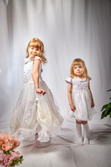 Fototapeta na wymiar Cute girls in beautifull dress with flowers in the studio on a white background. Young sisters posing indoors