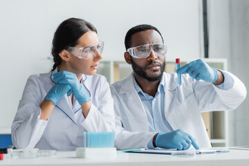 African american scientist in safety goggles holding test tube near colleague in latex gloves