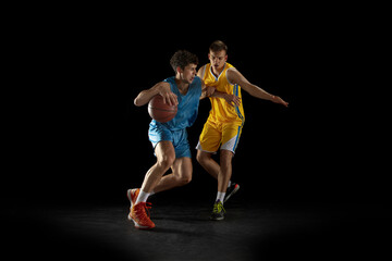 Fototapeta na wymiar Two basketball players in action and motion isolated on dark black studio background. Advertising concept. Strong Caucasian athletes practicing with basketball ball.