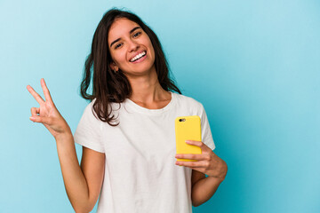 Young caucasian woman holding a mobile phone isolated on blue background joyful and carefree showing a peace symbol with fingers.