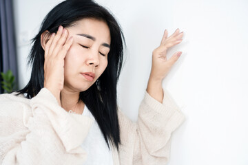 Asian woman having problem with Meniere's disease, fainting or dizziness hand holding her head leaning against the wall