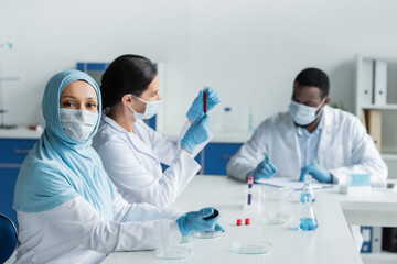 Muslim scientist in medical mask holding pipette near petri dishes and blurred colleagues in lab