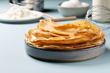 Stack of homemade crepes with ingredients for making thin pancakes on blue background.