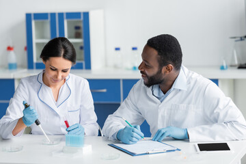 Fototapeta premium Smiling african american scientist in latex gloves writing on clipboard and looking at colleague with test tube