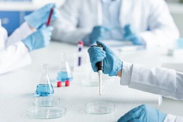Cropped view of scientist holding electronic pipette near petri dish and flasks