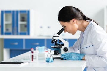 Side view of scientist in latex gloves using microscope near digital tablet with blank screen and...