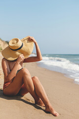Summer vacation. Beautiful stylish woman in hat sitting on sandy beach at sea waves, sunbathing and relaxing. Fit young female in swimsuit and straw hat enjoying vacation on tropical island