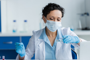 African american scientist in medical mask holding petri dish and pencil near laptop in laboratory