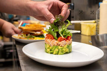 Chef's hands finishing assembling a salmon tartare with avocado and chopped cherry tomatoes on the metal kitchen table on a white tray