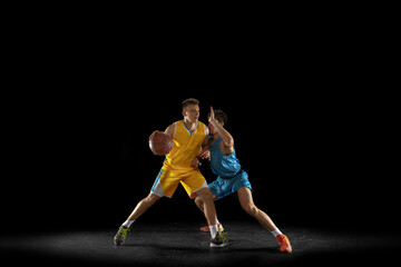 Obraz premium Two basketball players in action and motion isolated on dark black studio background. Advertising concept. Strong Caucasian athletes practicing with basketball ball.