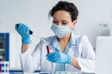 African american scientist in medical mask and latex gloves holding electronic pipette and test tube near blurred laptop