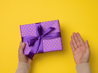two female hands are holding a purple gift box on a yellow background, happy birthday concept