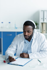 African american scientist in headphones using smartphone near petri dishes and clipboard