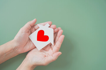 white house paper cut with red heart shape in hands on green background, homeless housing shelter...