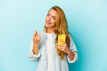Caucasian woman holding mobile phone isolated on blue background pointing with finger at you as if inviting come closer.