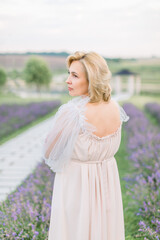 Back view of mature pretty blond woman in lavender field wearing light elegant long dress on cloudy summer day in the morning